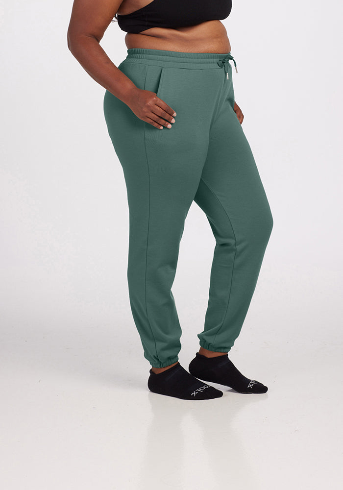 A person wearing a black crop top and Woolx's Parker Sweatpants in Duck Green, featuring temperature-regulating properties and a relaxed fit, stands against a white background, completing the look with black socks.