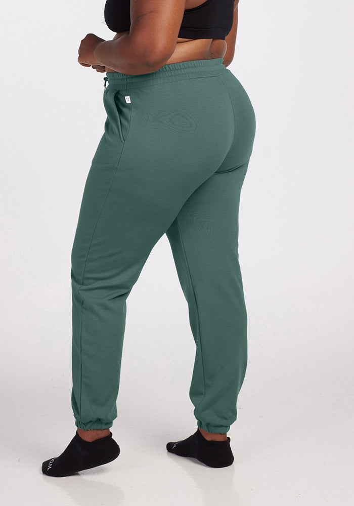 A person is wearing the Parker Sweatpants in Duck Green by Woolx, made from temperature-regulating Merino wool. They stand sideways, showcasing the side and back of their outfit, which also includes a black crop top and black socks. The background is plain white.