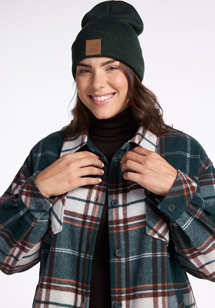 A person with shoulder-length brown hair is smiling while wearing a Woolx Baylor Beanie in Pine, a black turtleneck sweater, and a green and white plaid shirt. They are touching the lapels of their shirt with both hands. The background is plain white.