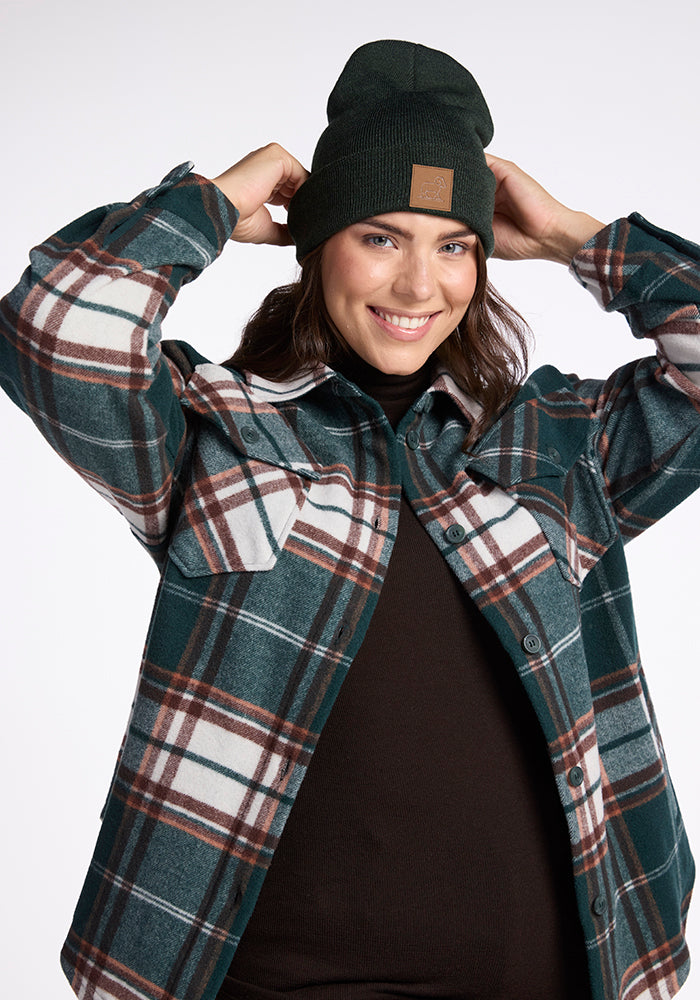 A person smiles while adjusting their green Woolx Baylor Beanie. They are wearing a dark brown shirt underneath a green, white, and brown plaid jacket. The background is plain and light-colored.