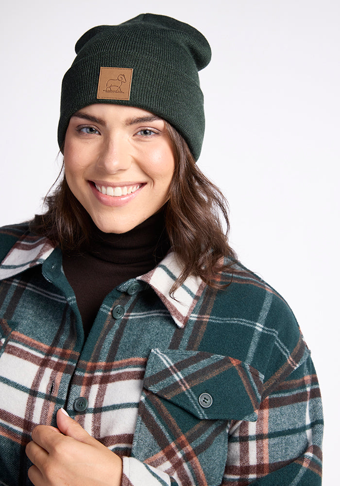 A person with shoulder-length brown hair is smiling while wearing a dark green Woolx Baylor Beanie featuring a brown patch, paired with a green, white, and brown plaid jacket over a black top. The background is plain and light-colored.