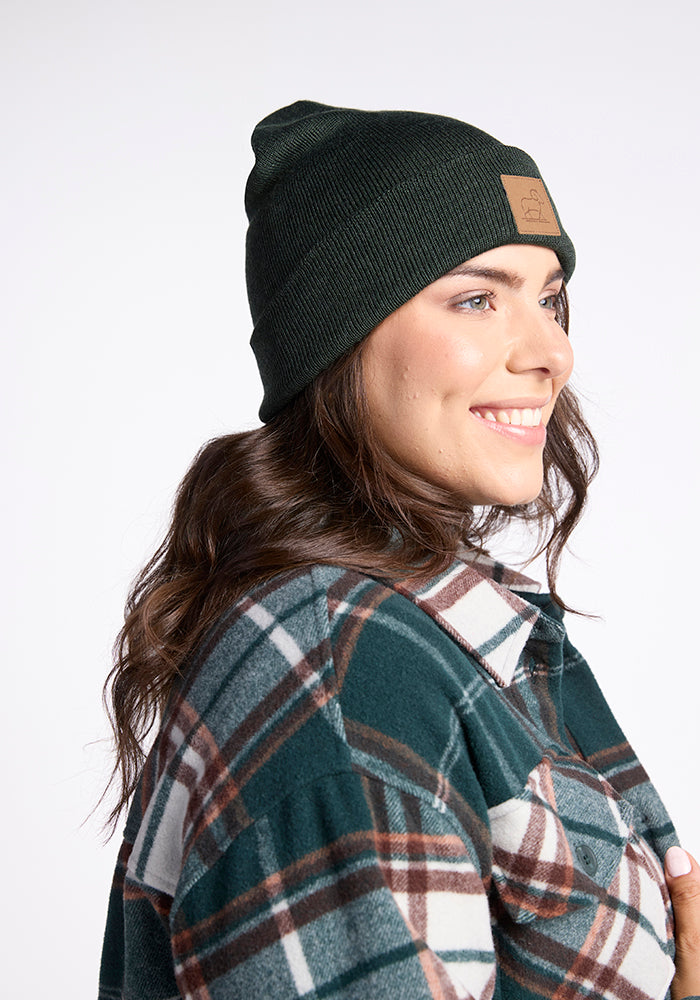 A person with long brown hair is smiling and wearing a dark green Baylor Beanie by Woolx along with a green, white, and brown plaid shirt. They are facing slightly to the left, clearly showcasing the beanie's logo on the front. The background is plain and well-lit.