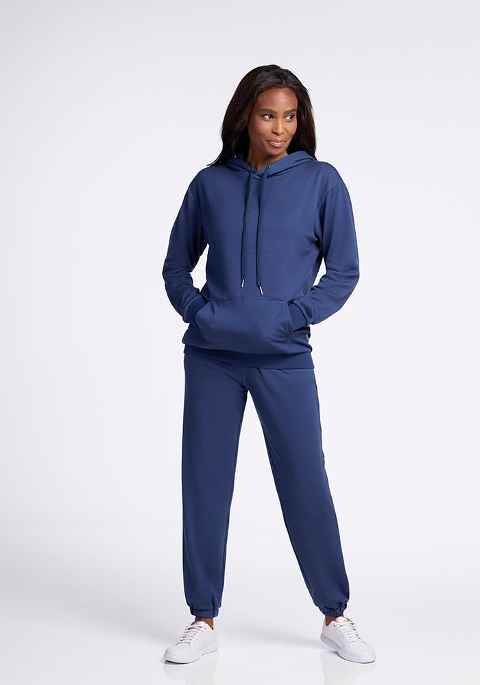 A person with long hair wearing a Woolx Avery Hoodie - Starry Night from the WoolAire Collection and matching jogger pants stands confidently. They have their hands in the hoodie's front pocket and are wearing white sneakers. The background is plain white.