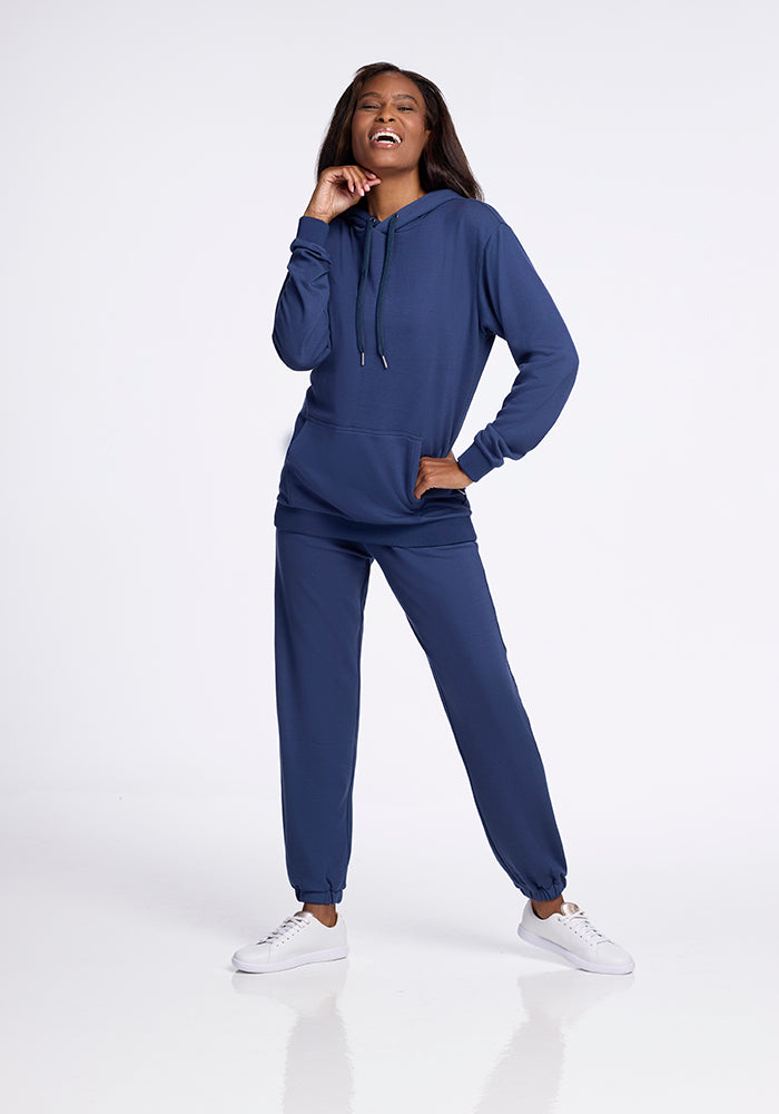 A person stands against a plain background, smiling and wearing a Woolx Avery Hoodie in Starry Night and matching joggers from the WoolAire Collection. They have one hand in the hoodie pocket and the other touching their chin. The cozy Merino wool outfit is completed with white sneakers.