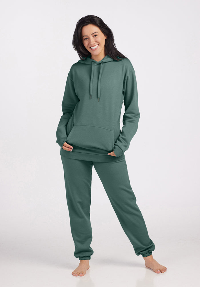 A person with long dark hair is smiling and standing barefoot in a relaxed pose. They are wearing matching duck green Avery hoodie and sweatpants from Woolx's WoolAire Collection™, crafted from soft Merino wool for ultimate comfort, against a plain white background.