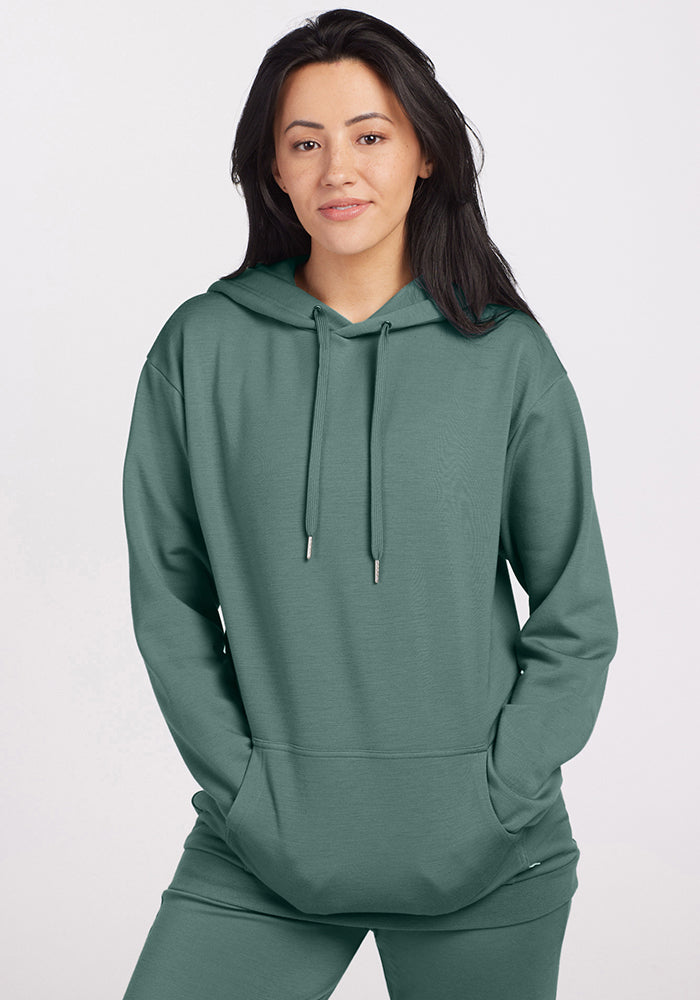 A person wearing a Duck Green Avery Hoodie from Woolx gazes at the camera with a relaxed expression. Their hoodie, made from soft Merino wool, includes a kangaroo pocket. They have long dark hair, and the background is plain and light-colored. 