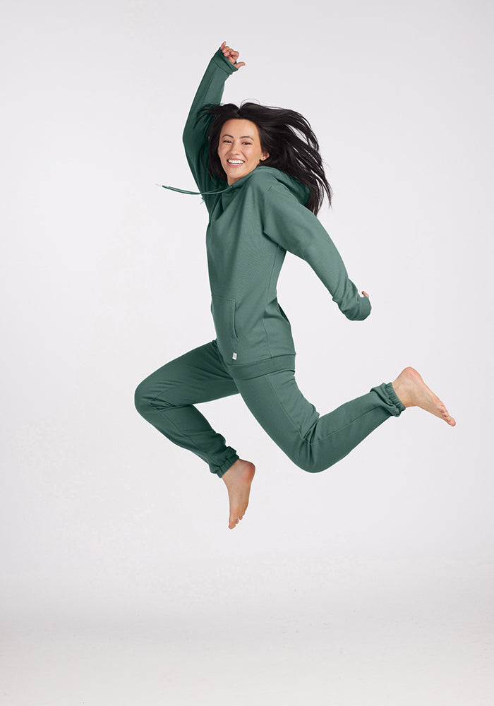 A person wearing a Duck Green Avery Hoodie from the Woolx brand is joyfully jumping in the air against a plain background, with arms raised, feet off the ground, and smiling widely.