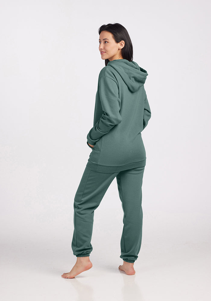 A person stands barefoot on a plain background, enveloped in the gentle comfort of a Woolx Avery Hoodie in Duck Green from the WoolAire Collection™, paired with matching green jogger pants, slightly turning away from the camera.