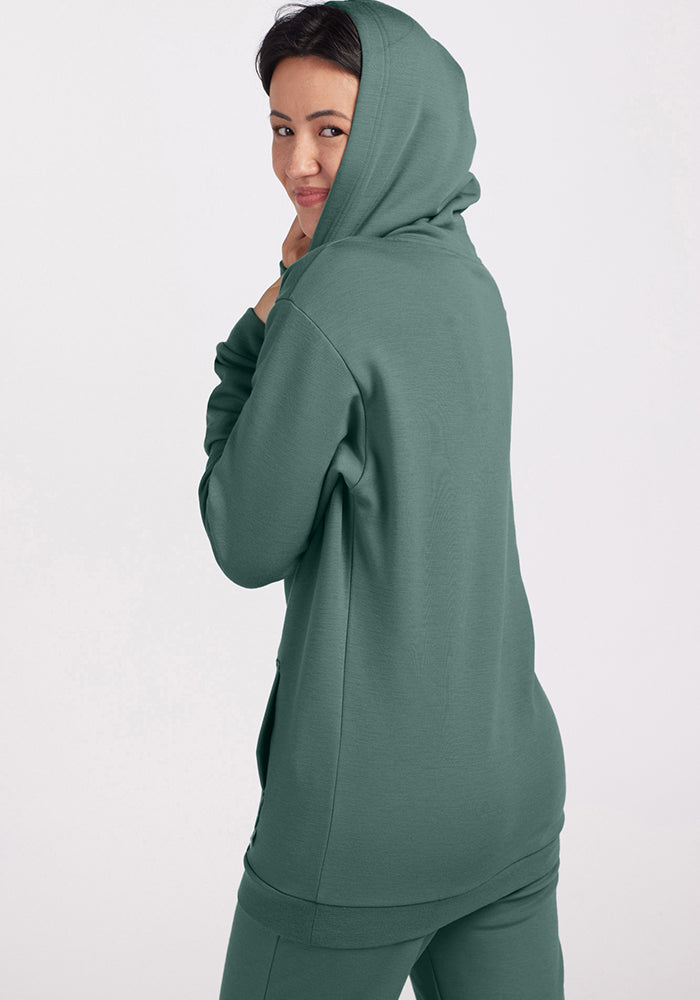 Dressed in a Duck Green Avery Hoodie from Woolx, made of luxurious Merino wool with the hood up and matching pants, the person turns slightly to look over their shoulder with a smile. The plain white background accentuates the cozy and relaxed ensemble.