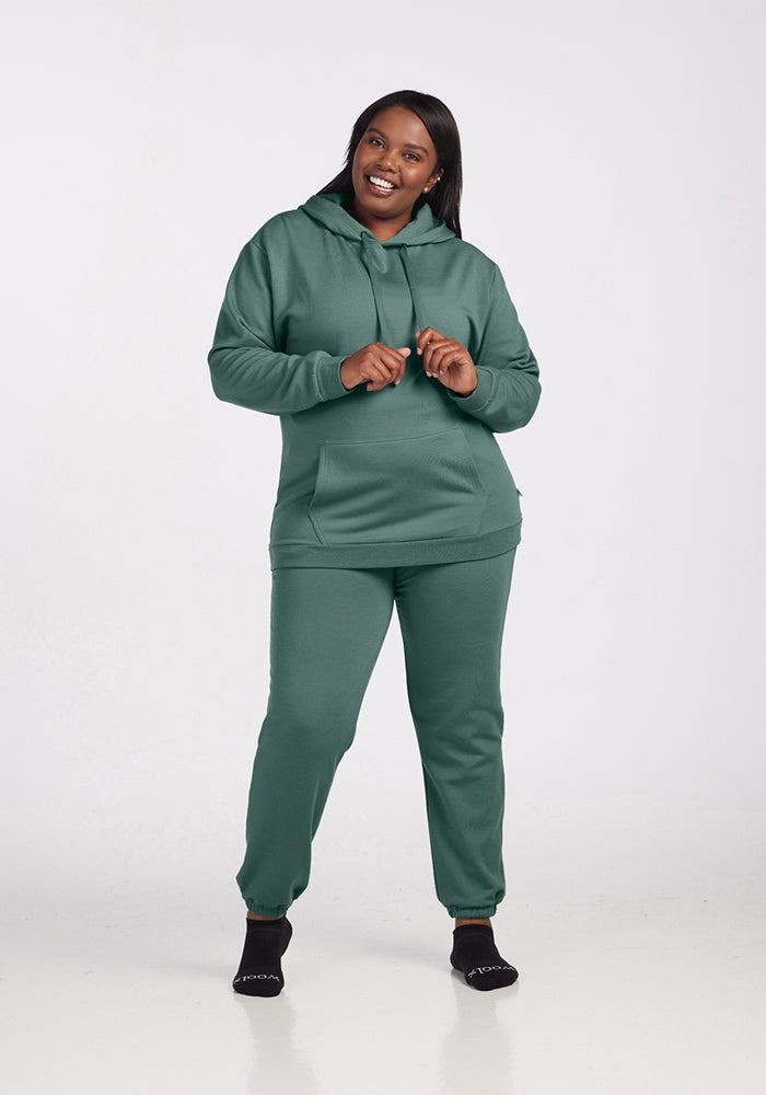 A person wearing a Woolx Avery Hoodie in Duck Green and coordinating pants from the WoolAire Collection™ smiles as they stand against a plain white background. They hold the drawstrings of the hoodie, crafted from soft Merino wool, and wear black socks.