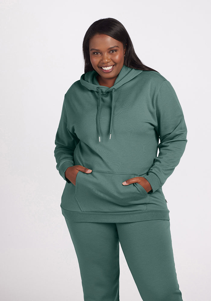 A person smiles while wearing the Avery Hoodie - Duck Green from Woolx, paired with matching pants. Crafted from soft Merino wool, the green hoodie features a front pocket and drawstrings against a plain white background.