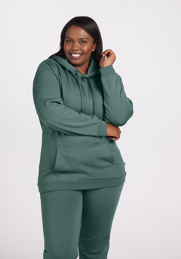 A person smiling while wearing the Avery Hoodie - Duck Green and matching pants from Woolx. Crafted in comfortable Merino wool, they stand in a relaxed pose against a plain background.