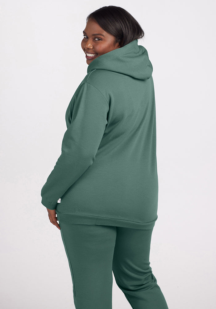 A person wearing an Avery Hoodie in Duck Green by Woolx, paired with matching sweatpants from the WoolAire Collection™, smiles while standing sideways. The outfit, crafted from soft Merino wool, stands out against the plain white background.