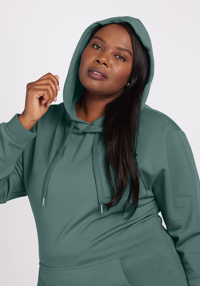 Dressed in a Duck Green Avery Hoodie from Woolx, they strike a confident pose with the hood up and drawstrings hanging down. Their long, dark hair perfectly frames a face that meets the camera's gaze against a simple background.