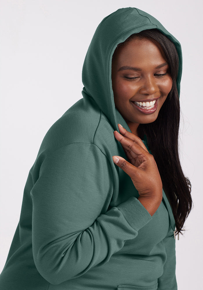 A woman smiling warmly, enveloped in the cozy comfort of a Duck Green Avery Hoodie from Woolx, with her hand gently resting on her shoulder.