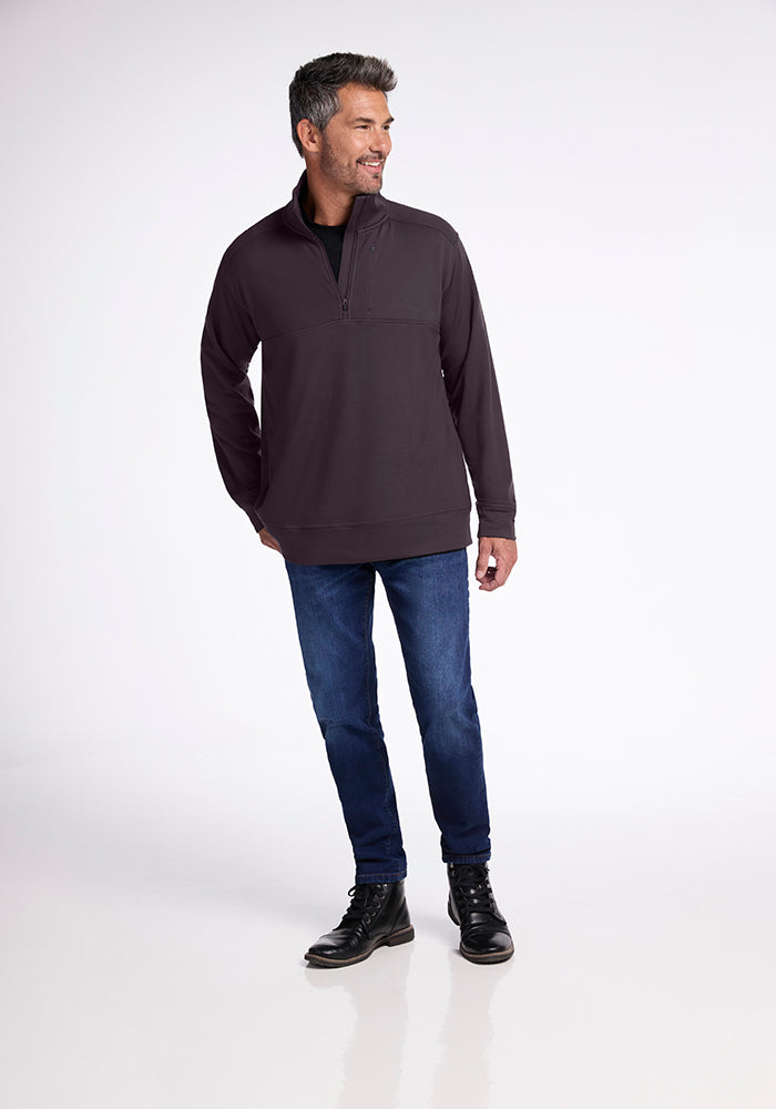 A man dressed in the Woolx Wool Aire™ Mason - Chocolate Plum, a dark midweight half-zip pullover crafted from Australian Merino Wool, pairs it with blue jeans and black boots. He stands against a plain white background, smiling and looking to the side with his hands in his pockets.