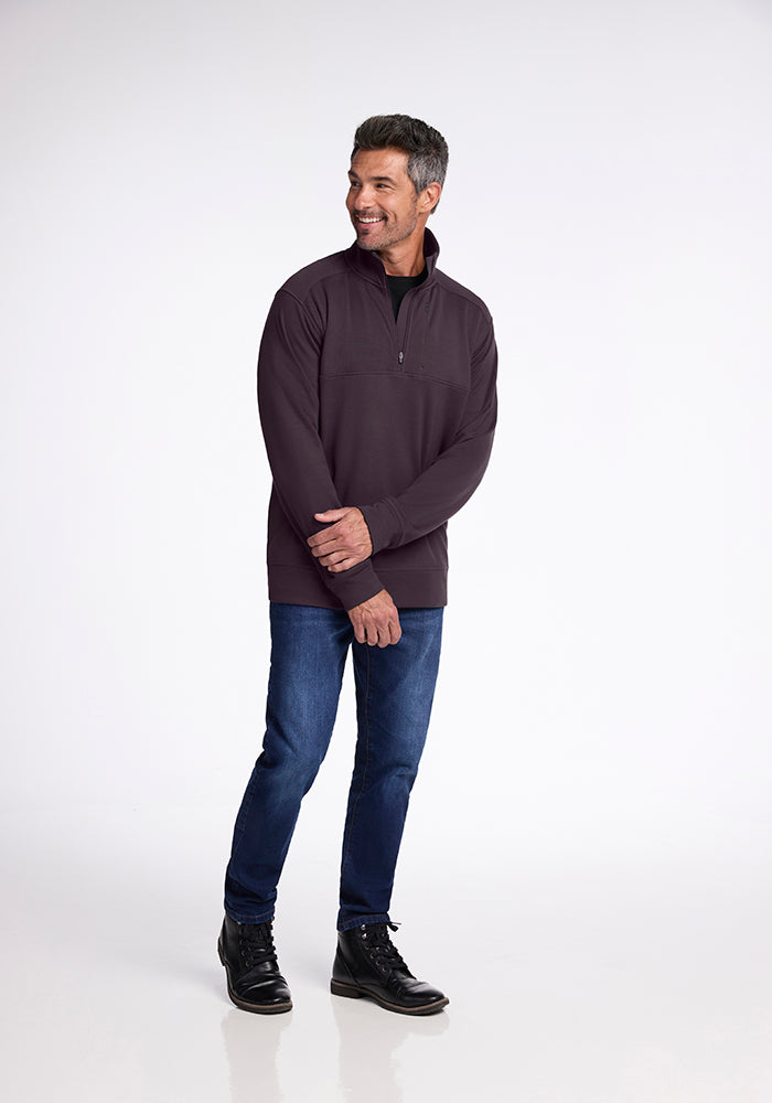 A man wearing the Woolx Wool Aire™ Mason sweater in Chocolate Plum, along with blue jeans and black boots, stands against a plain white background. He is smiling and looking to the side with his hands clasped in front of him.