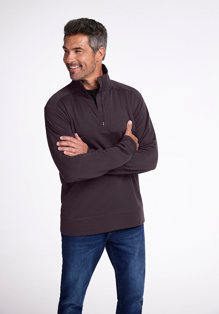 A man smiles while standing with his arms crossed, wearing a Woolx Wool Aire™ Mason in Chocolate Plum over a black shirt and blue jeans. The midweight design offers comfort and style against the plain, light-colored background.