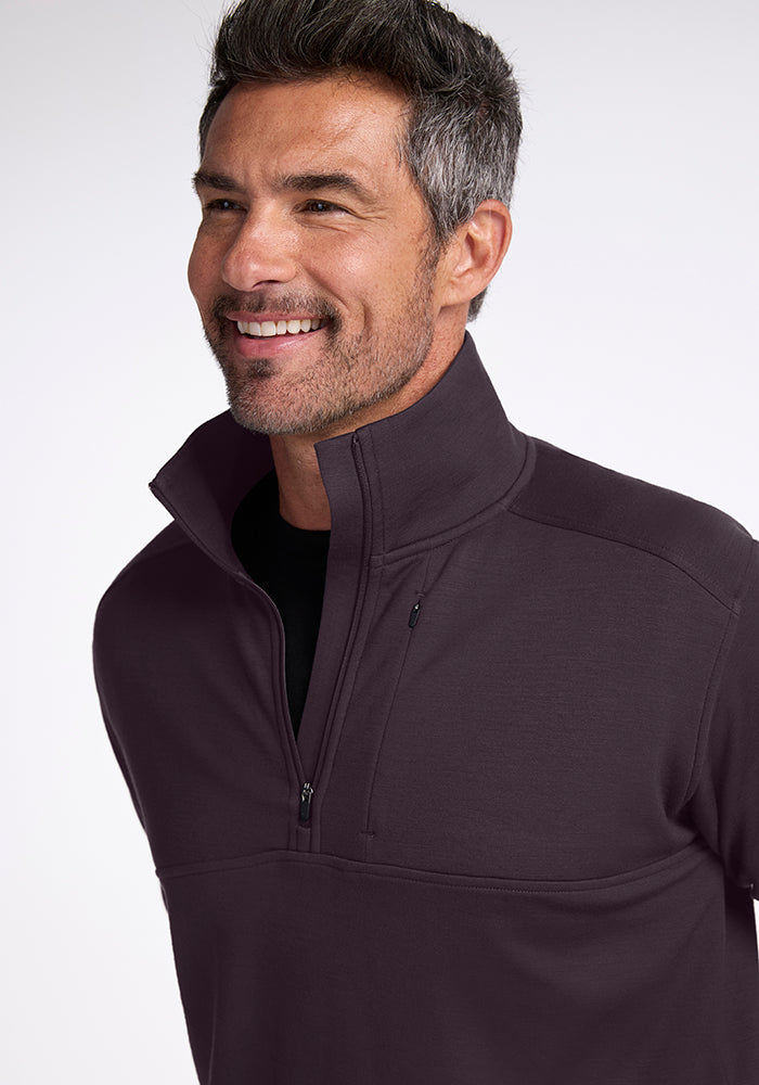 A man with short hair and a beard smiles while wearing the Woolx Wool Aire™ Mason pullover in Chocolate Plum, a dark midweight half-zip sweater made of Merino Wool. The background is plain and light-colored.