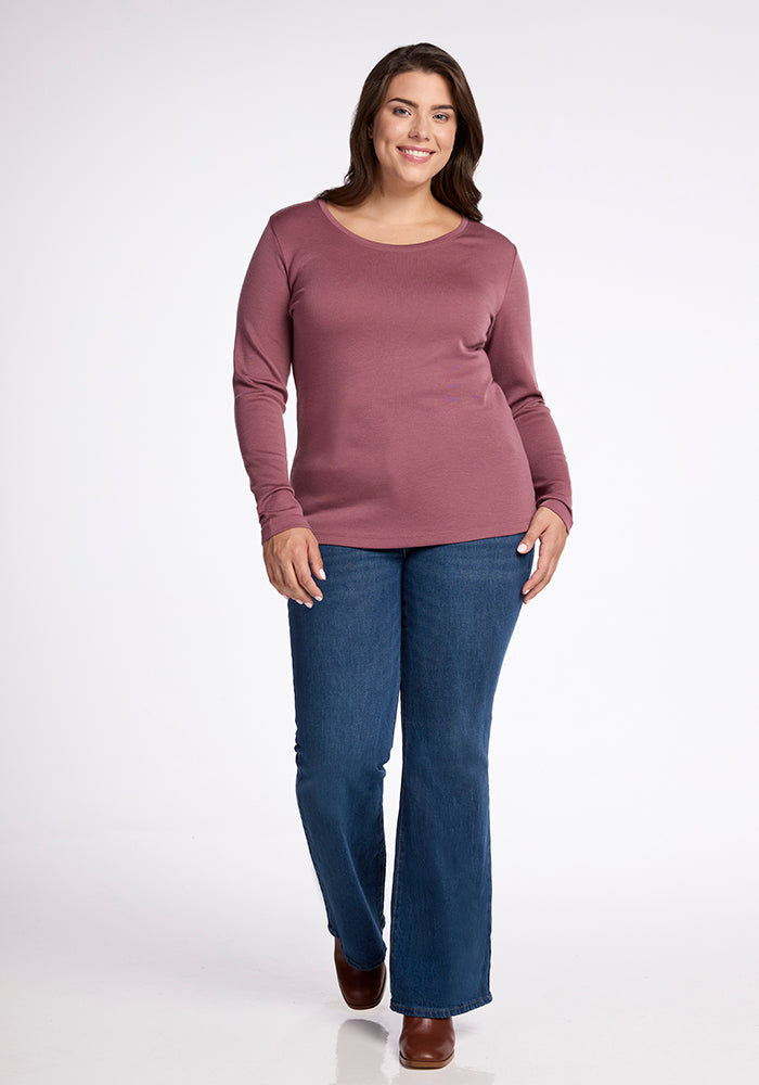 A person is standing and smiling at the camera. They are wearing a long-sleeved, maroon Kenzie Scoop Neck top from Woolx, blue jeans, and brown shoes. The background is plain white.