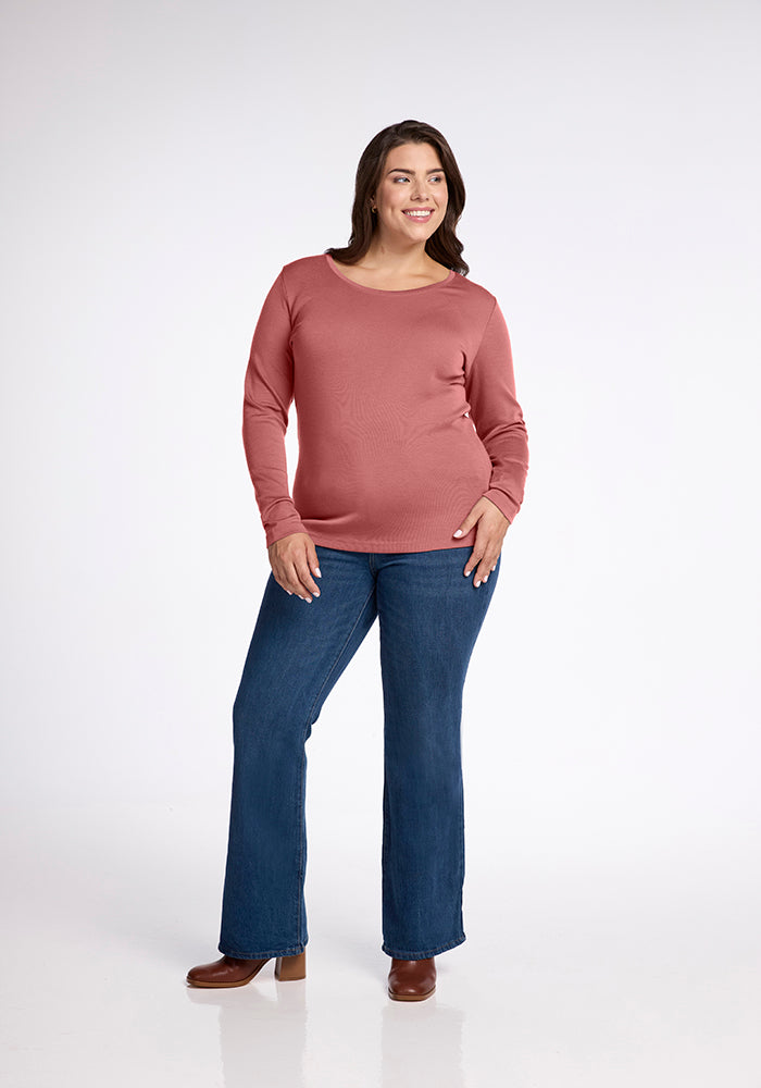 A person stands smiling against a plain, light-colored background. They are wearing the Woolx Kenzie Scoop Neck, a long-sleeved, rose-colored Merino wool top paired with blue jeans and brown shoes—an ensemble that's as stylish as it is comfortable for three seasons. Their hands are relaxed, one resting on their hip and the other hanging by their side.