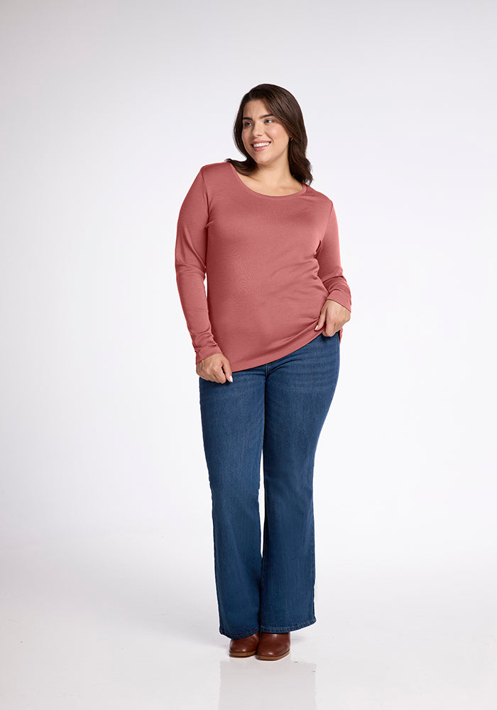 A woman with long brown hair is wearing a versatile, long-sleeve Kenzie Scoop Neck from Woolx in rose color and blue jeans. She is standing and smiling, with her left hand in her pocket and her right hand resting on her thigh. She is wearing brown shoes against a plain white background, embodying business casual style.