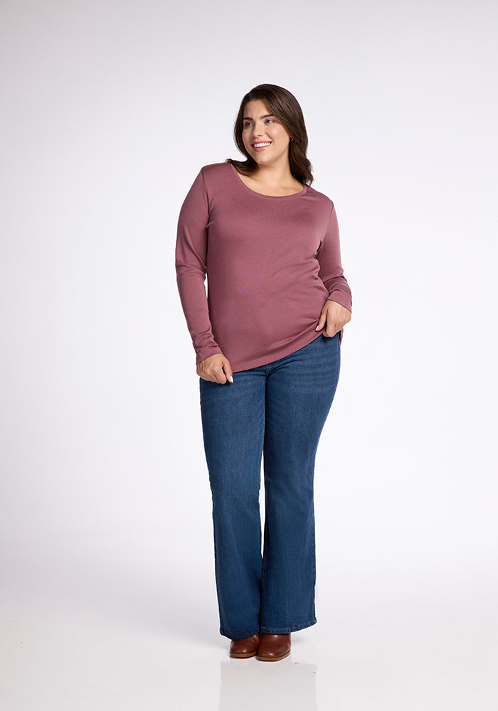 A woman with long brown hair is standing and smiling, dressed in a long-sleeved Kenzie Scoop Neck shirt by Woolx, paired with blue flared jeans and brown ankle boots—perfect for versatile apparel. The background is a plain, light grey color.