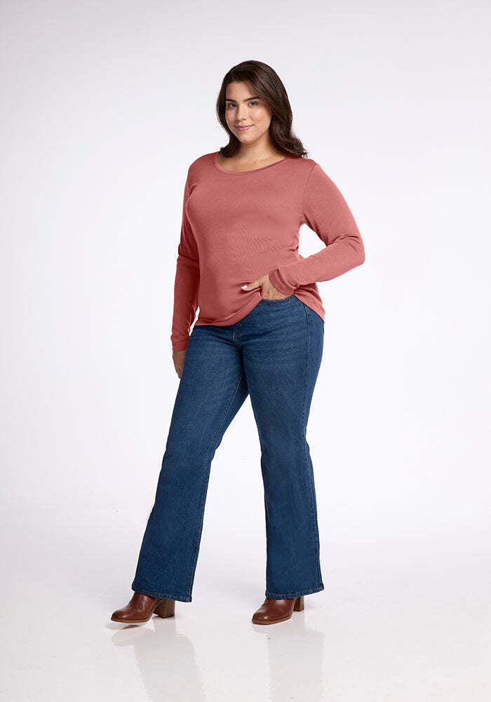 A woman with long dark hair poses in front of a plain white background, smiling. She is wearing a long-sleeved, pink Kenzie Scoop Neck shirt from Woolx and blue jeans with brown, heeled ankle boots. One hand rests on her hip while the other is down by her side.