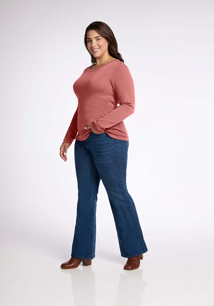 A woman is standing and facing slightly to the left, smiling at the camera. She is wearing a versatile, long-sleeved Kenzie Scoop Neck from Woolx paired with blue jeans and brown shoes. The background is plain white.