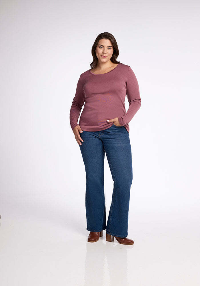 A woman stands against a plain white background, wearing the Kenzie Scoop Neck from Woolx and a pair of blue jeans. She is smiling with her left hand in her pocket and sporting brown heeled shoes. Her outfit exemplifies versatile apparel perfect for business casual wear.