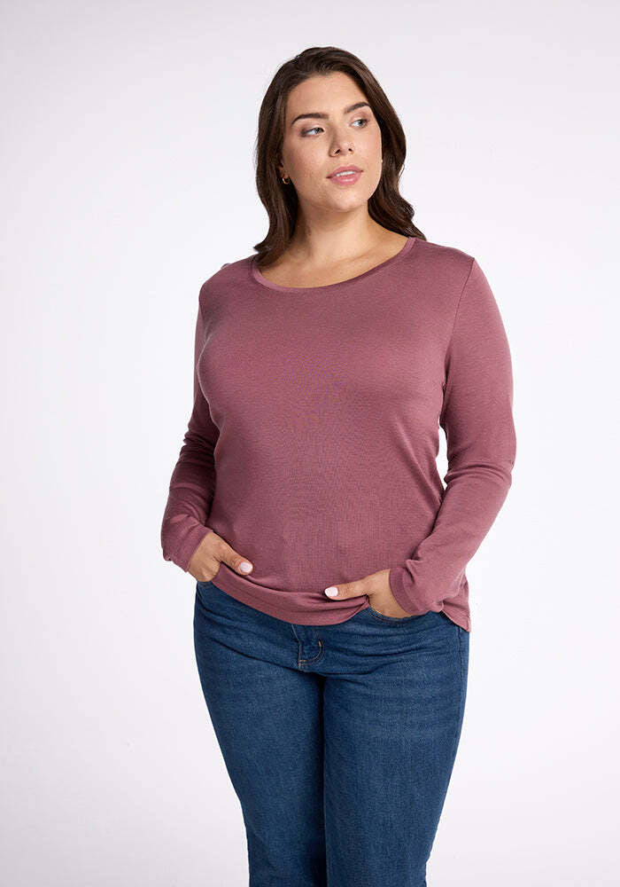 A woman with long, wavy hair wears a mauve, long-sleeve Kenzie Scoop Neck top by Woolx and blue jeans. She has her hands in her pockets and looks off to the side against a plain, light-colored background. 