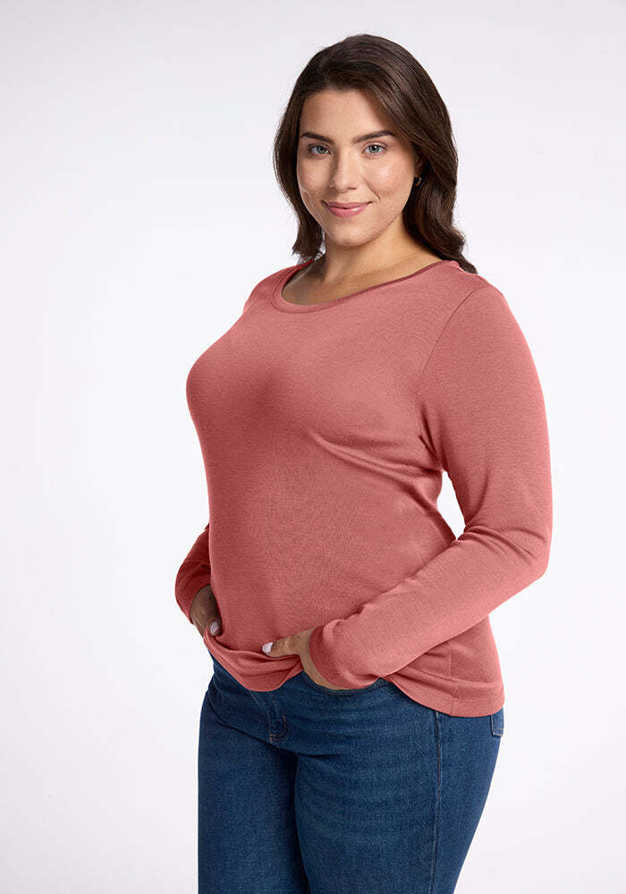 A woman with long dark hair is smiling and facing the camera. She is wearing a comfy, long-sleeved Kenzie Scoop Neck by Woolx in a pinkish-red color and blue jeans. Her hands are in the pockets of her jeans, and she is standing against a plain white background.