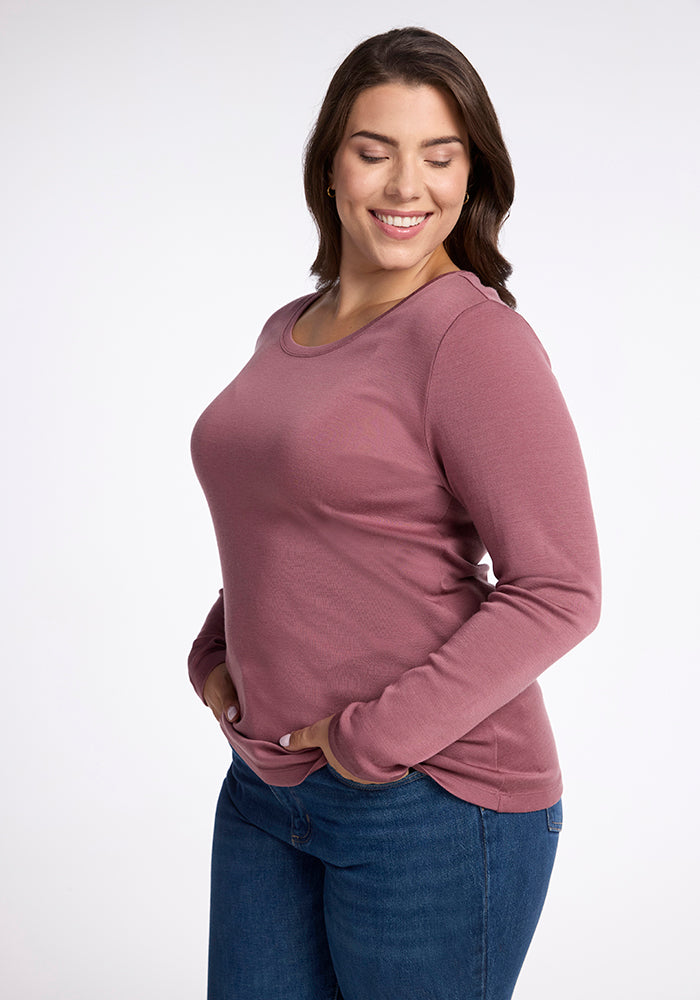 A person with long hair is smiling with their eyes closed, standing with their hands in the pockets of their blue jeans. They are wearing a long-sleeved, dusty pink Kenzie Scoop Neck shirt made of comfy Merino wool by Woolx. The background is plain white.