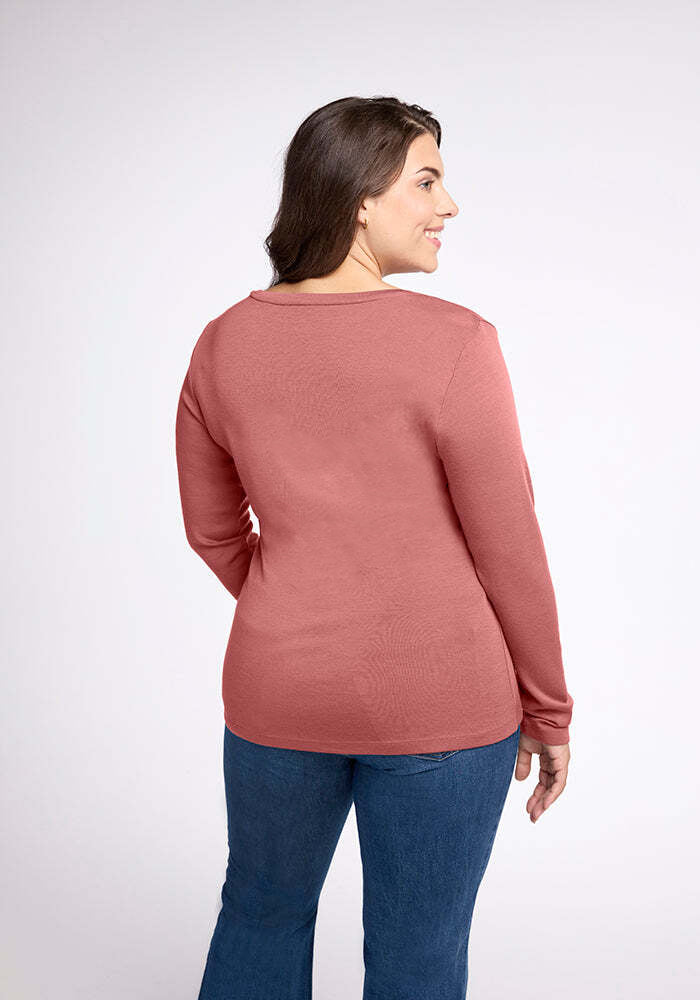 A person with long hair, wearing a Woolx Kenzie Scoop Neck Merino wool shirt in rose and blue jeans, is standing and facing away from the camera, looking over their left shoulder with a slight smile. The background is plain and light-colored.