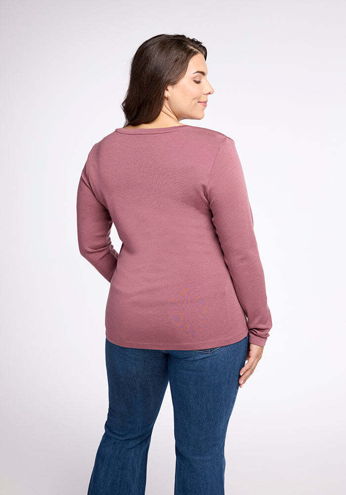 A person with long hair stands with their back to the camera, wearing a versatile Woolx Kenzie Scoop Neck in a long-sleeved mauve/rose-colored style and blue jeans. The plain white background emphasizes the comfy shirt.