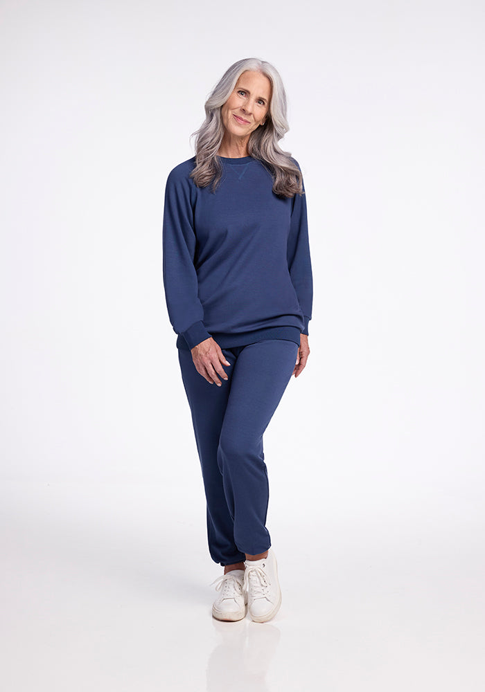 A woman with long gray hair stands against a plain white background, smiling and wearing the matching Bailey Crewneck Sweatshirt - Starry Night and sweatpants from Woolx, paired with white sneakers. She exudes a relaxed and casual appearance, showcasing the comfort of Australian Merino Wool.