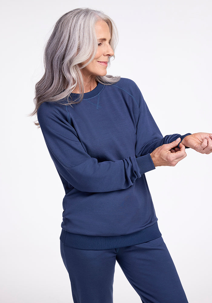 A woman with long, gray hair stands in a relaxed pose wearing a matching navy blue Bailey Crewneck Sweatshirt in Starry Night and pants from the Woolx Wool Aire Collection. She looks down and adjusts her sleeve, with a slight smile on her face. The background is plain white.