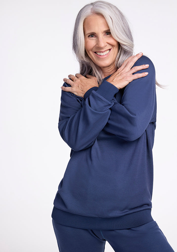 An older woman with long gray hair smiles while embracing herself in a cozy pose, wearing a Bailey Crewneck Sweatshirt - Starry Night from Woolx. Her navy blue sweater and matching pants complement the plain white background perfectly.