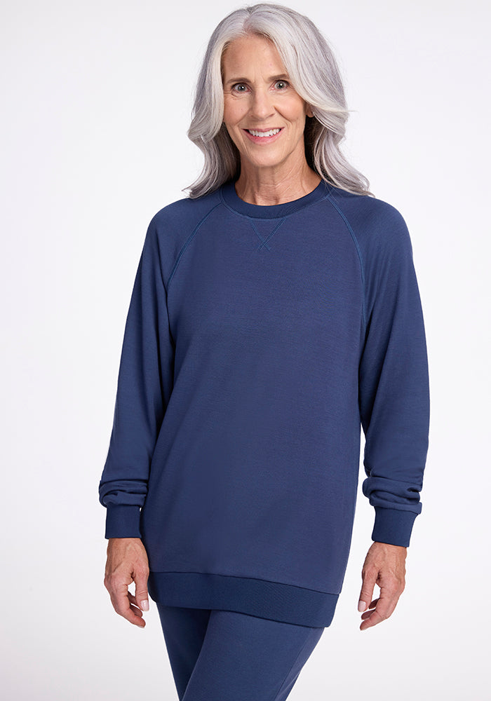 A woman with long gray hair smiles at the camera. She is wearing a Starry Night Bailey Crewneck Sweatshirt from Woolx and matching pants from the Wool Aire Collection. The background is plain white.