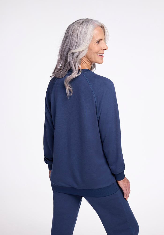 An elderly woman with long, wavy gray hair is standing and smiling, facing slightly away from the camera. She is wearing a matching navy blue Bailey Crewneck Sweatshirt in the Starry Night color by Woolx and pants set made from sustainable clothing materials. The background is plain and light-colored.