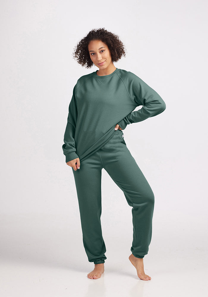 A person with curly hair stands barefoot, wearing a matching green outfit from the Woolx collection. The sustainable Bailey Crewneck Sweatshirt and sweatpants, crafted from Merino Wool in Duck Green, complement their pose with one hand on their hip against a plain white background.