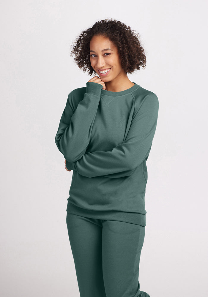 A person with curly hair smiles while standing with one hand under their chin, showcasing the Bailey Crewneck Sweatshirt in Duck Green from the Woolx collection. They are wearing a green Merino wool outfit against a plain white background.