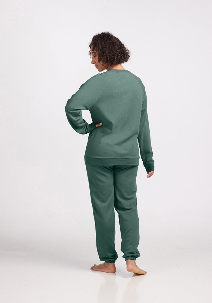 A person with curly hair stands barefoot on a plain white background, showcasing the Wool Aire Collection. Dressed in the Bailey Crewneck Sweatshirt - Duck Green by Woolx and matching pants made from Merino wool, the person faces away from the camera, looking over their left shoulder.