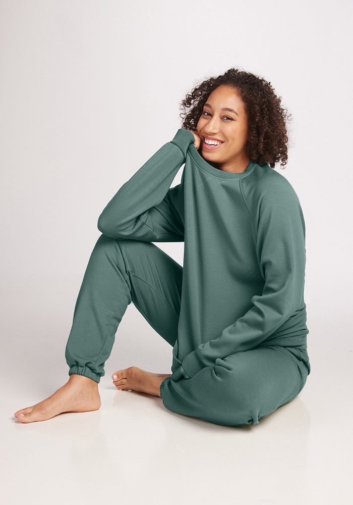 A person sits on the floor in a cozy, sustainable Bailey Crewneck Sweatshirt in Duck Green from Woolx, paired with matching green sweatpants, smiling with one knee up. The Merino Wool outfit complements the simple, light-colored setting beautifully.