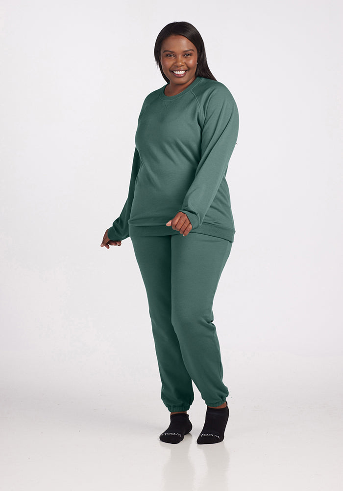 A person smiling and posing in the Duck Green Bailey Crewneck Sweatshirt from Woolx with matching pants. They are wearing black socks and standing against a plain white background.