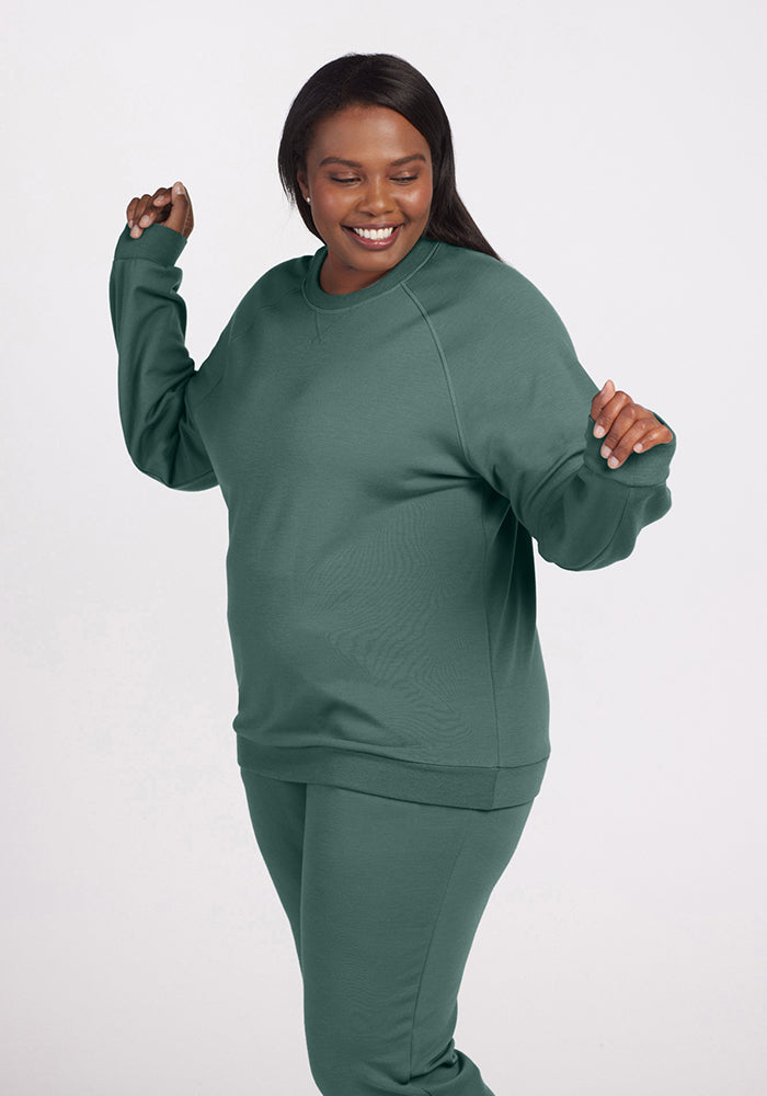 A person is smiling and posing in a relaxed stance, wearing a coordinated set from Woolx featuring the Bailey Crewneck Sweatshirt in Duck Green with merino wool pants. They are standing against a plain white background.