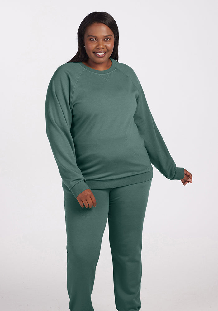 A person smiling while wearing the Bailey Crewneck Sweatshirt in Duck Green from Woolx, along with matching sweatpants. They are standing against a plain, light-colored background.