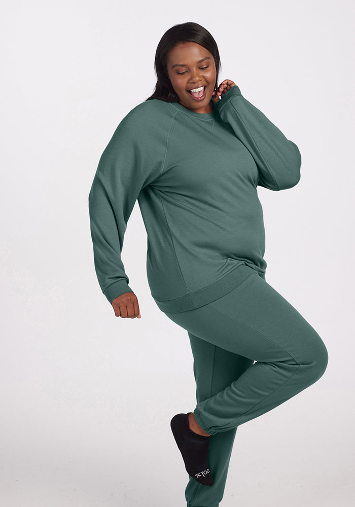 A person joyfully poses in a Duck Green Bailey Crewneck Sweatshirt from Woolx, playfully raising one leg while smiling with closed eyes. Wearing dark slippers, they seem to be relishing a cheerful moment against a plain background, celebrating the comfort of Merino wool.