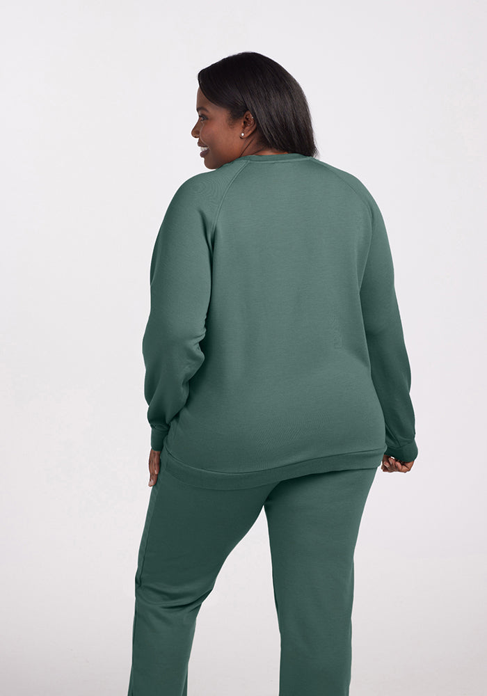 A person is standing and facing away from the camera, dressed in a matching dark green ensemble featuring the Bailey Crewneck Sweatshirt in Duck Green by Woolx. The eco-friendly sweatshirt and pants accentuate the elegance of Merino wool against an off-white background.