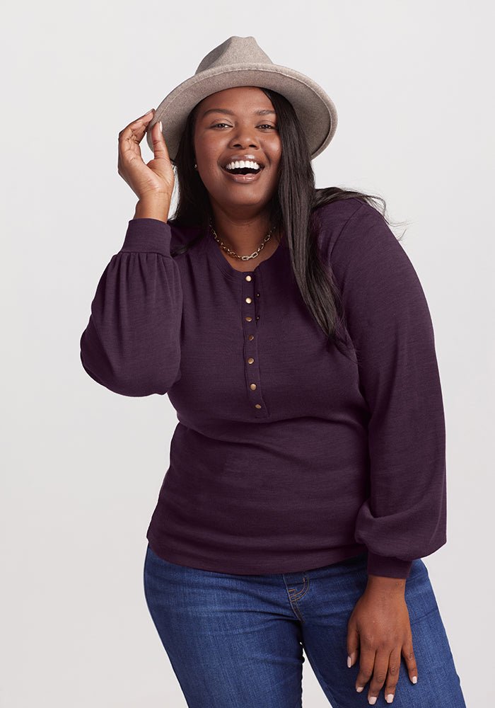 A person smiles while wearing a gray hat, the Charlotte Puff Sleeve Top in Deep Plum by Woolx with Henley-style front buttons, and blue jeans. They hold the brim of the hat with one hand against a plain, light-colored background.